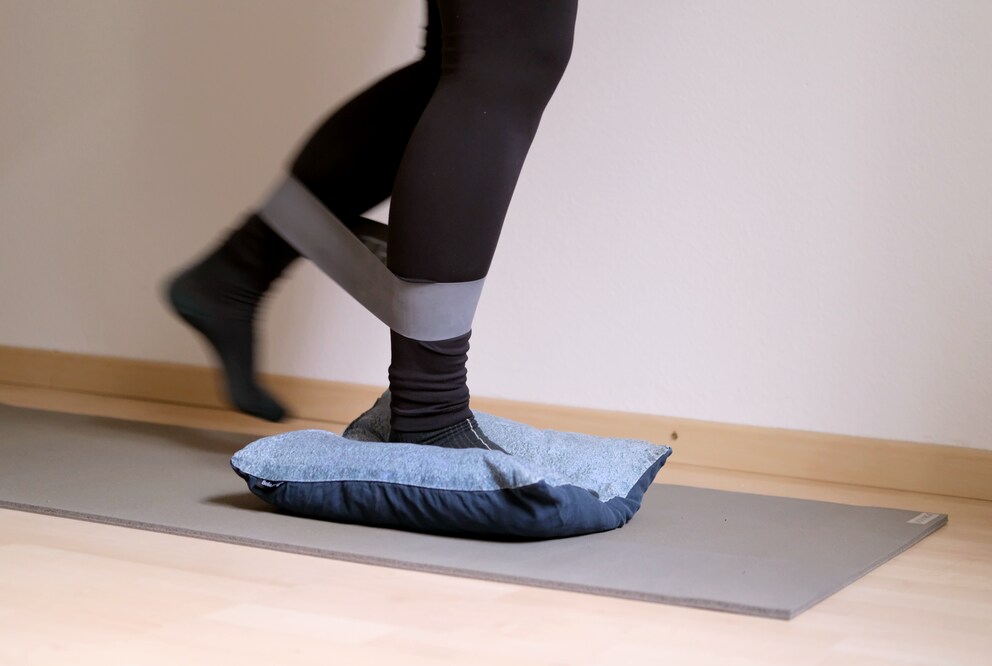 Währenddem das eine Bein auf dem Kissen steht, wird das andere im Halbkreis von vorne nach hinten gegen den Widerstand des Fitnessbands bewegt.